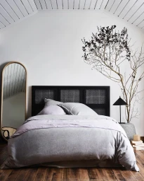 Minimalist bedroom interior with a black cane headboard, grey linen bedding, arched floor mirror, and a dried decorative branch.
