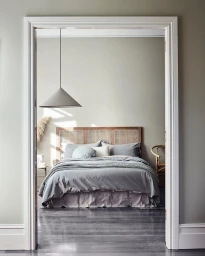 Interior view through a white doorway showing a made bed with grey linens and a cane headboard in a minimalist bedroom.