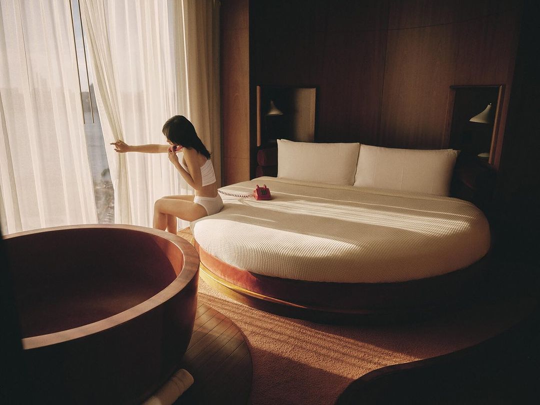 Woman sits on round bed near window in luxury hotel room with vintage red phone and wood tub; warm lighting and elegant decor.