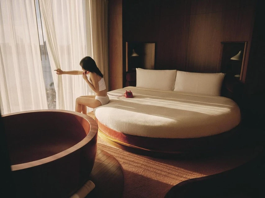 Woman sits on round bed near window in luxury hotel room with vintage red phone and wood tub; warm lighting and elegant decor.