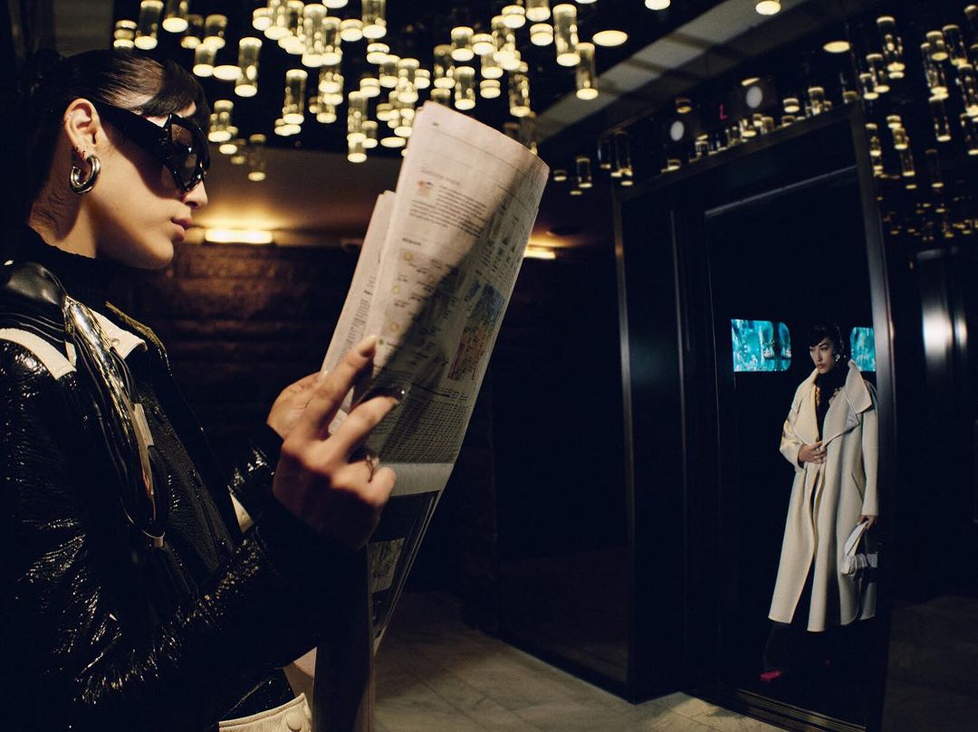 Woman reading newspaper in stylish outfit in front of elevator with monitors and contemporary chandelier in dark interior setting.