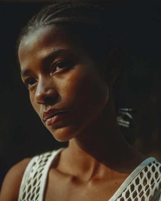 Close-up studio portrait of a woman with cornrow braids, wearing a white mesh top, and bathed in soft natural light creating dark shadows.