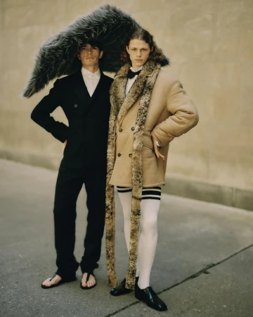 Two men pose together outdoors; one in a black suit with a large fur accessory, the other in a beige coat with fur trim.