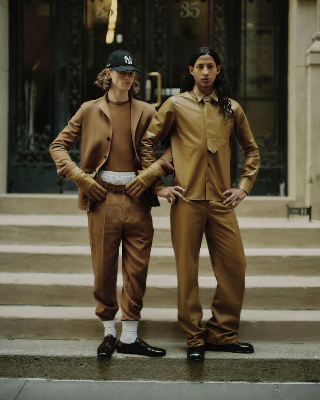 Two young men pose on stairs outdoors in matching brown outfits, one wearing a baseball cap, in an editorial fashion shot.