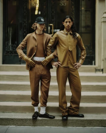 Two young men pose on stairs outdoors in matching brown outfits, one wearing a baseball cap, in an editorial fashion shot.