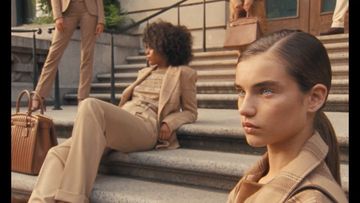 Models in matching tan suits pose on outdoor stone steps with purses. Fashion editorial shot featuring neutral colors and urban background.