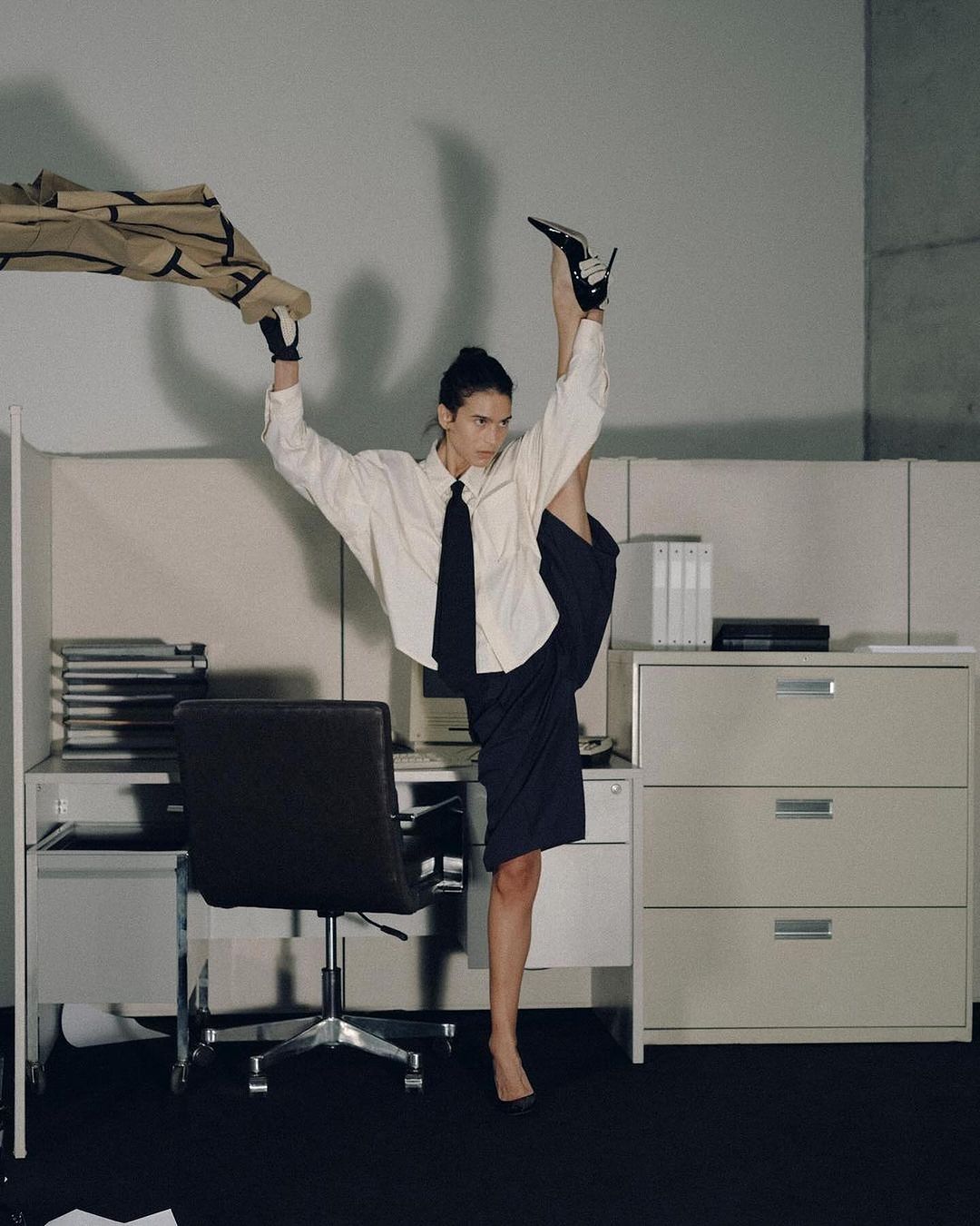 A woman in an office is standing in a split with one leg straight up and one leg on the ground next to a desk with office supplies.