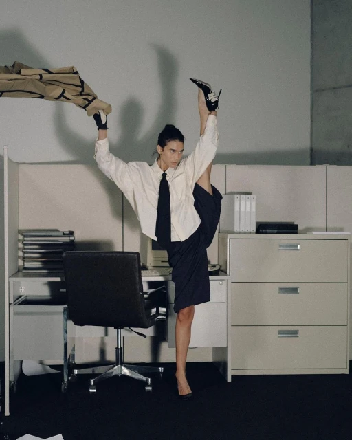 A woman in an office is standing in a split with one leg straight up and one leg on the ground next to a desk with office supplies.