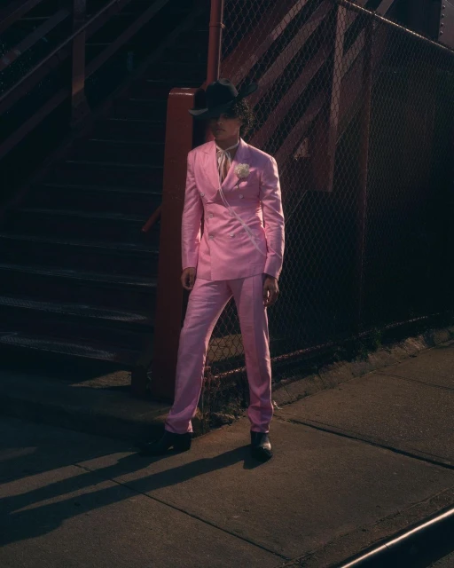 Man in pink suit and black hat stands on a sidewalk near a metal structure, creating long shadows in the evening light.