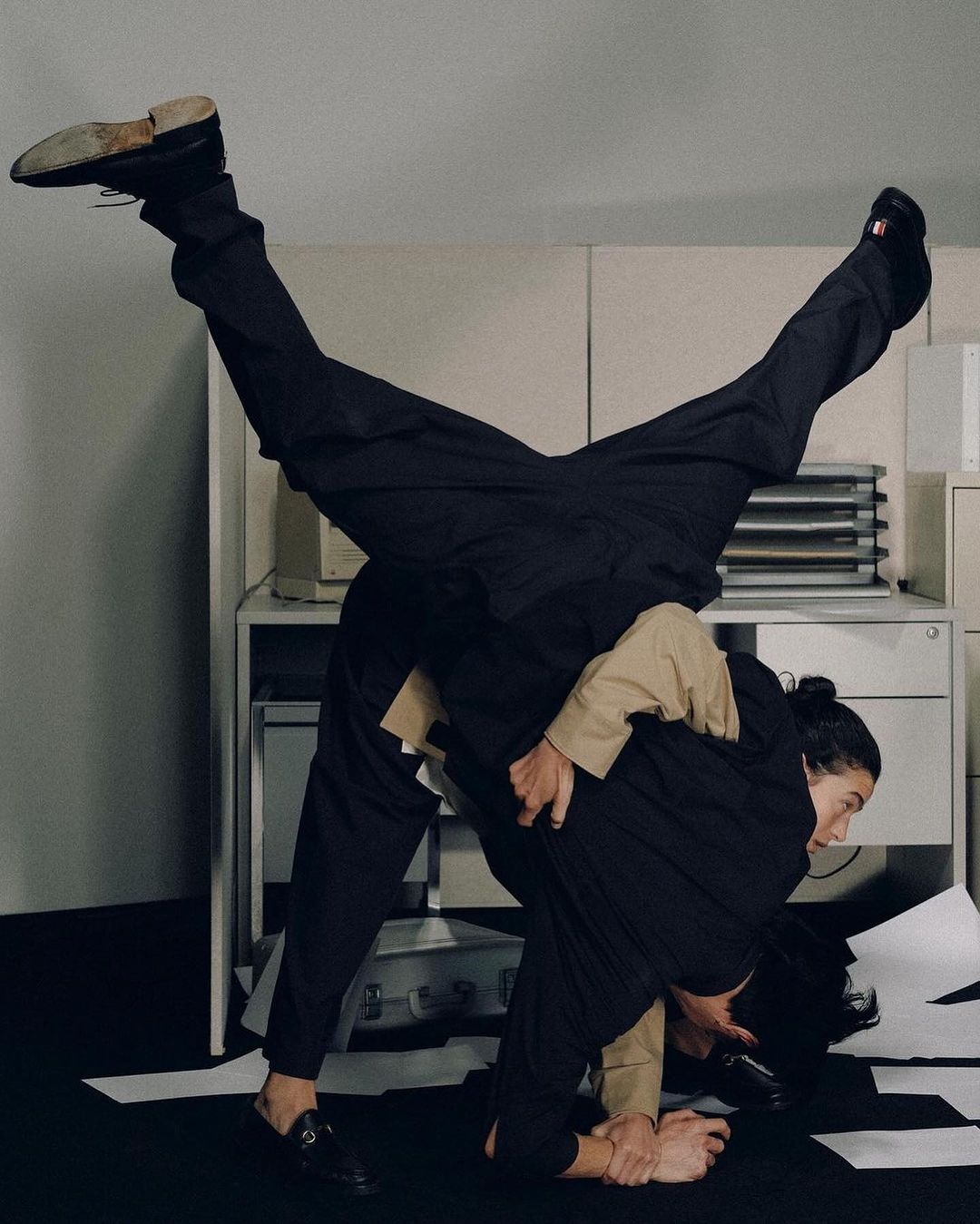 Two adult men perform an acrobatic pose in front of office cabinets. One man supports the other who is upside down with legs extended.