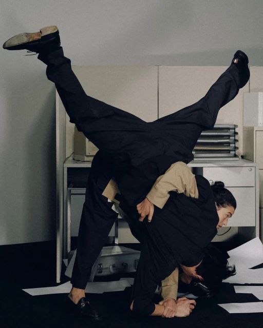 Two adult men perform an acrobatic pose in front of office cabinets. One man supports the other who is upside down with legs extended.