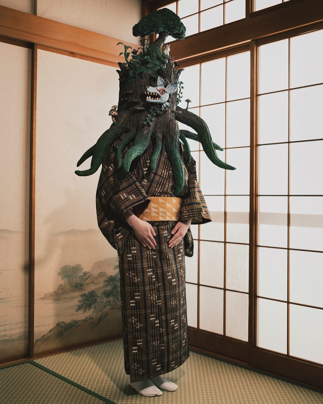 Person in unique tree-inspired costume, complete with mask and kimono, standing indoors with shoji screen, creating an editorial, artsy image.