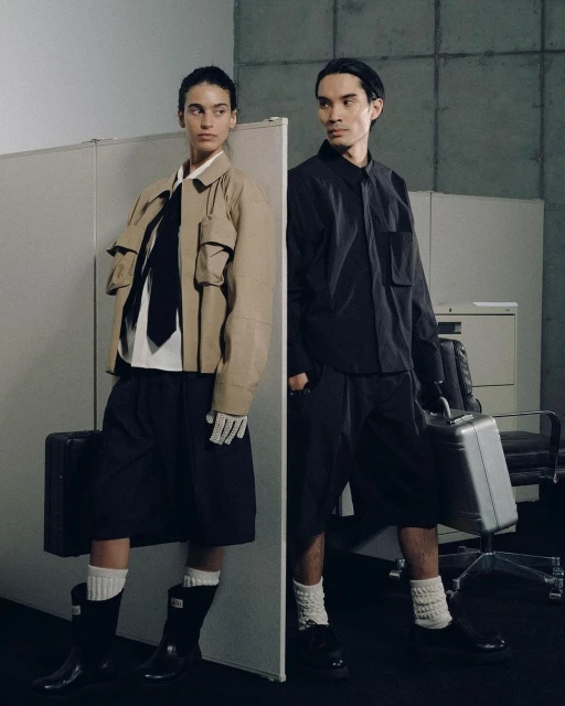 Two people in black and tan outfits pose in an office setting with a light grey wall and desk. One holds a briefcase.