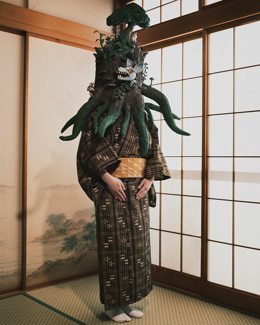Person in elaborate tree costume with fox mask standing in front of shoji screen in room with tatami mat flooring.