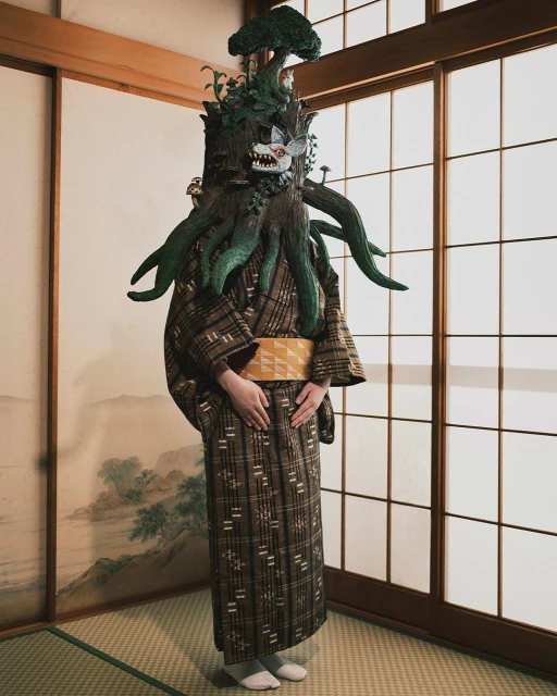 Person in elaborate tree costume with fox mask standing in front of shoji screen in room with tatami mat flooring.