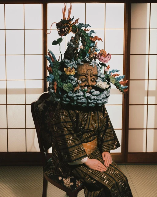 Person seated wearing a kimono and an elaborate headpiece featuring a dragon, flowers, and a mask, in front of a shoji screen.