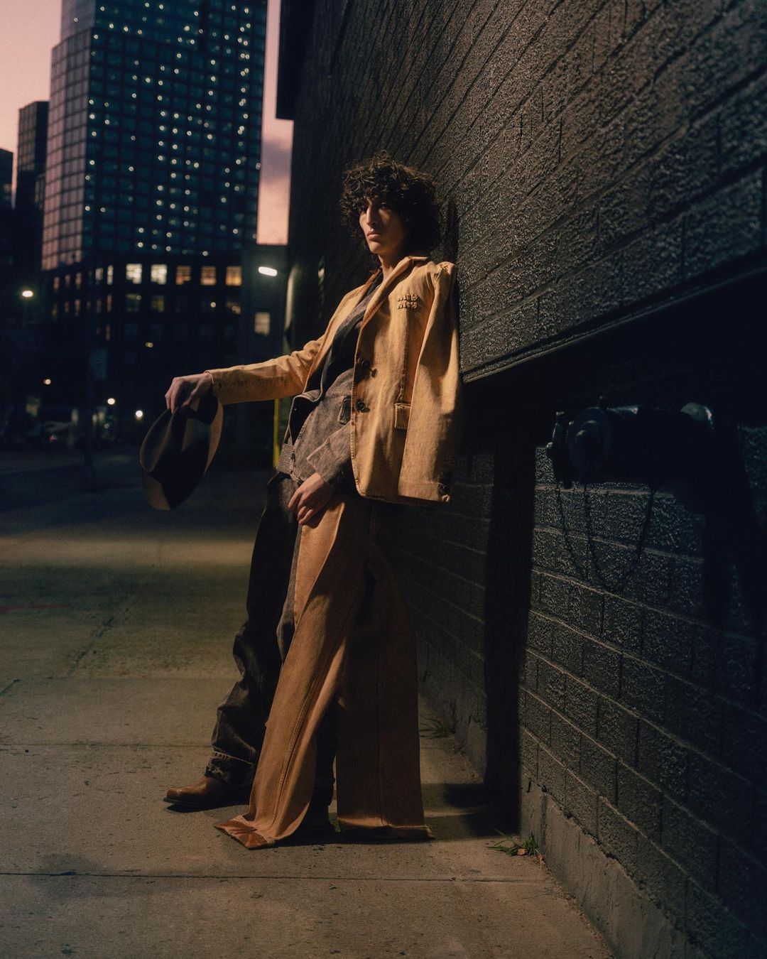 Woman in yellow suit leans against brick wall at night. City buildings and street scene in background with dim, ambient lighting.