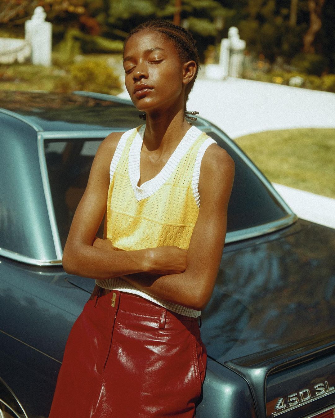 Woman in yellow knit top and red leather pants poses against a vintage blue car outdoors in natural light with trees in background.