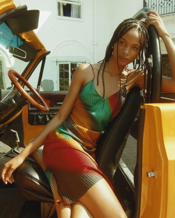 Young Black woman in a multicoloured patterned slip dress seated inside a vintage yellow convertible car.