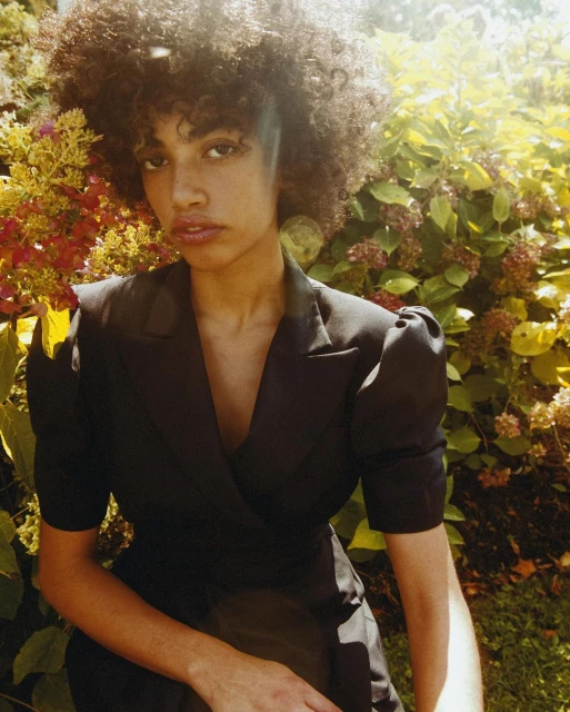 Portrait of a young woman with afro hairstyle wearing a black jacket, posing outdoors with natural lighting and leafy plant background.