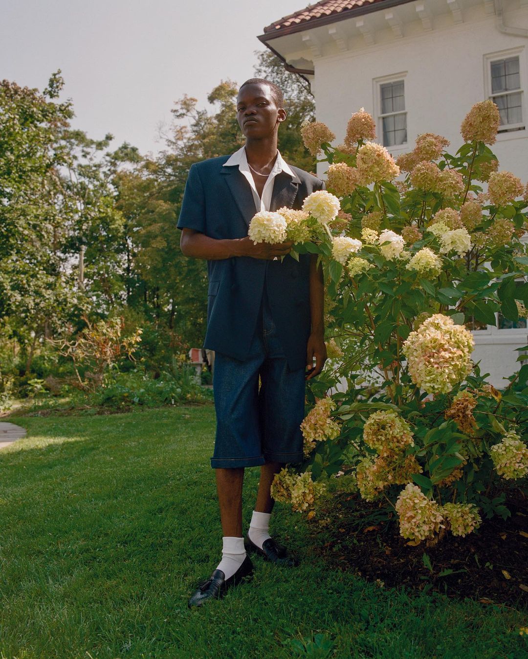 Adult male posing outdoors in blazer and shorts, holding white flowers in a garden with a house and trees in the background.