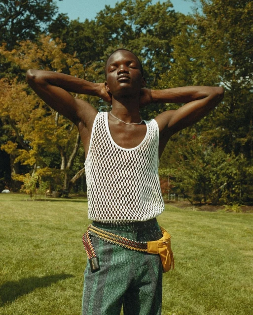 An adult Black man stands with hands behind his head in a white mesh tank top and green pants outdoors with grass and trees in the background.
