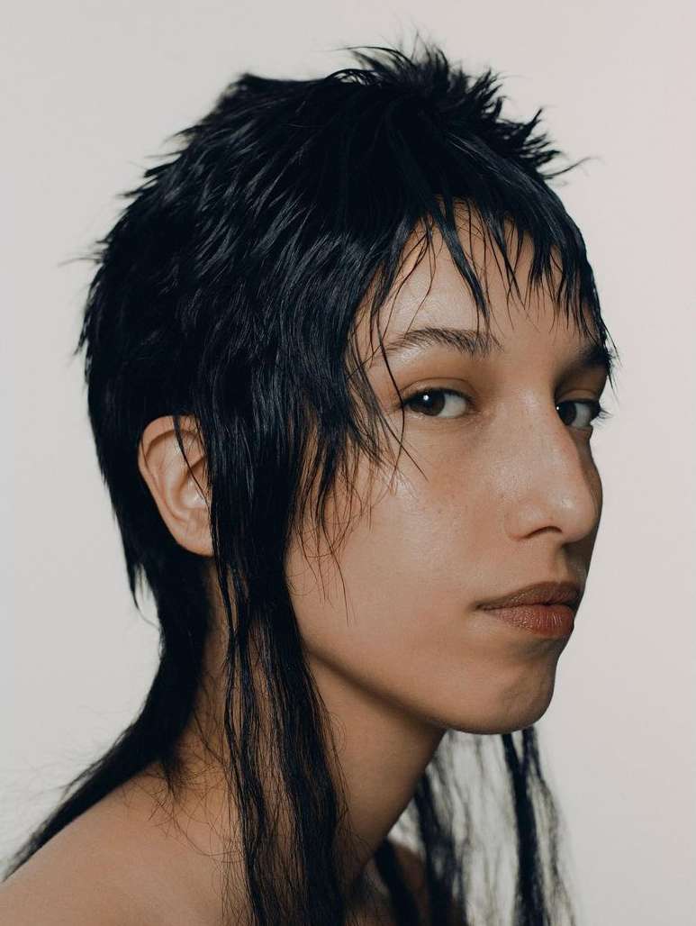 Portrait of a person with short, dark, textured hair and light skin in a close-up shot against a bright, neutral background.