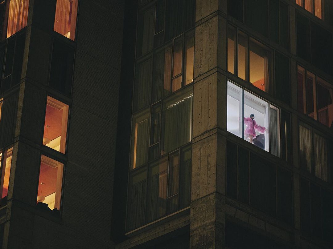 Night view of a high-rise apartment building, a woman visible in a brightly lit window wearing a pink robe.