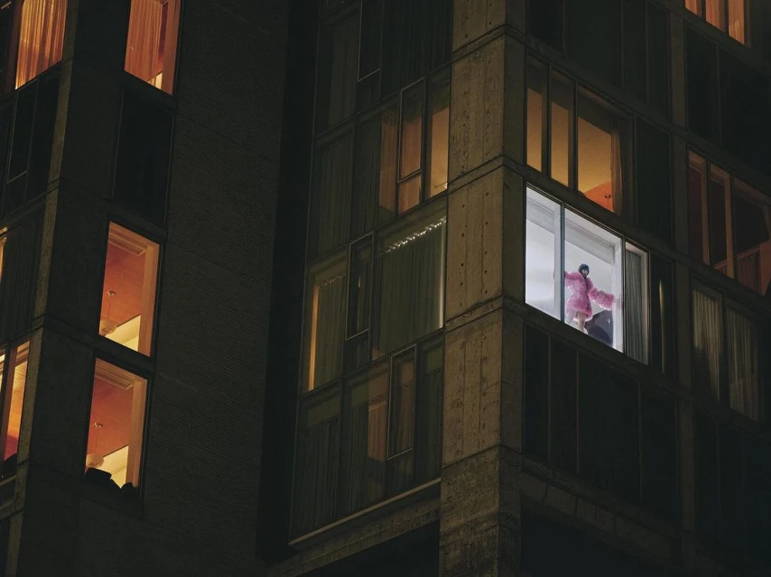 Night view of a high-rise apartment building, a woman visible in a brightly lit window wearing a pink robe.