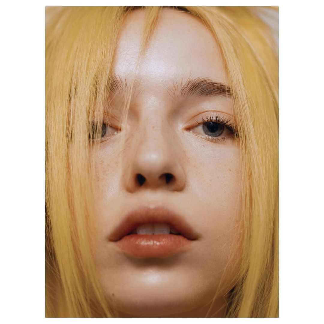 Close-up portrait of a young fair-skinned woman with light blonde hair and blue eyes, facing the camera, minimalist composition, high-key lighting.