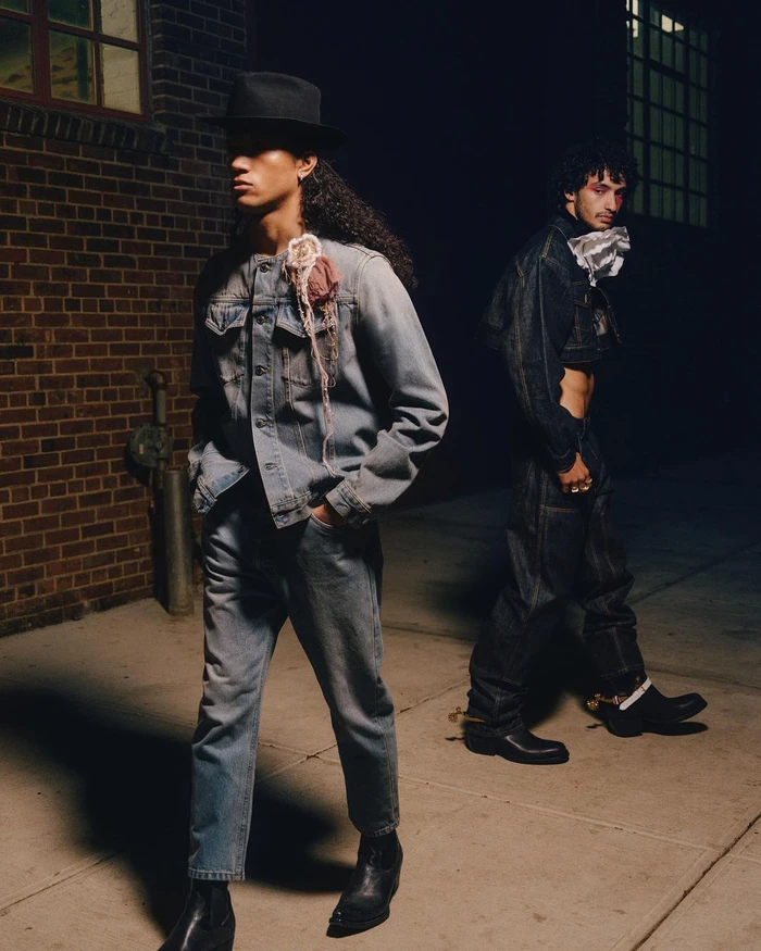 Two men walking in denim outfits at night on an alleyway pavement with brick walls behind them.