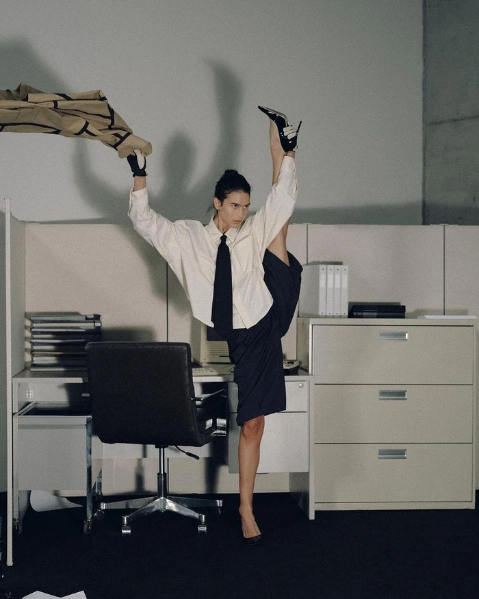 Model in white shirt and dark skirt holding leg high in an office by Bon Duke