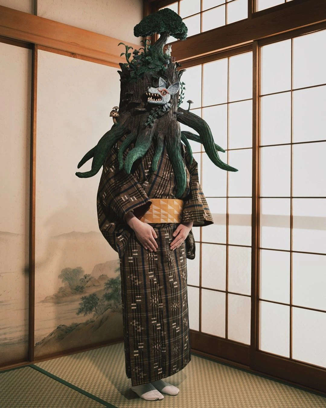 Person wearing an elaborate tree stump headpiece with a wolf mask and traditional patterned kimono in a Japanese room.