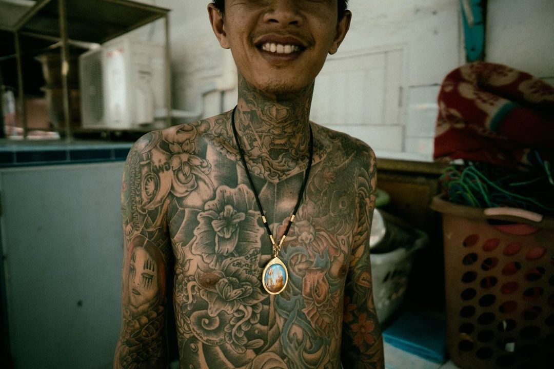 Smiling man with extensive tattoos covering his torso, neck, and arms, wearing a pendant necklace, indoors.