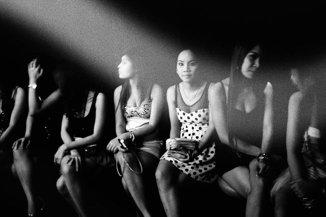 Several young adult women are sitting together in a dark room, partially illuminated by a ray of light in this black and white image.