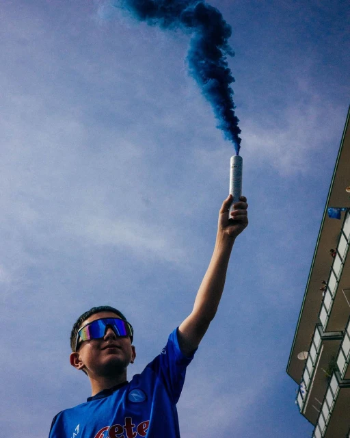 A person holds a blue smoke canister against a blue sky, wearing sunglasses and a sports jersey with a building in the background.