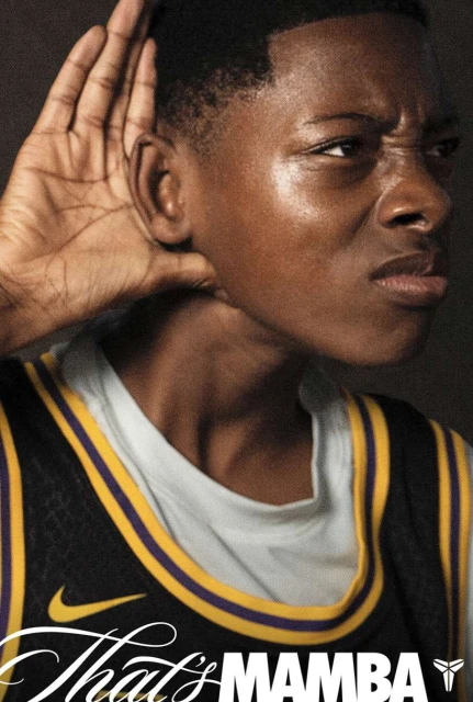 Young man in a basketball jersey puts a hand to his ear, listening intently, with an inquisitive facial expression against a dark backdrop.