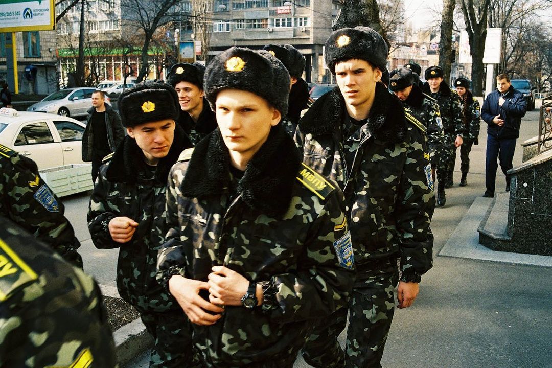 Group of people in military camouflage uniforms and winter hats walk on a city street near buildings and parked cars on a sunny day.