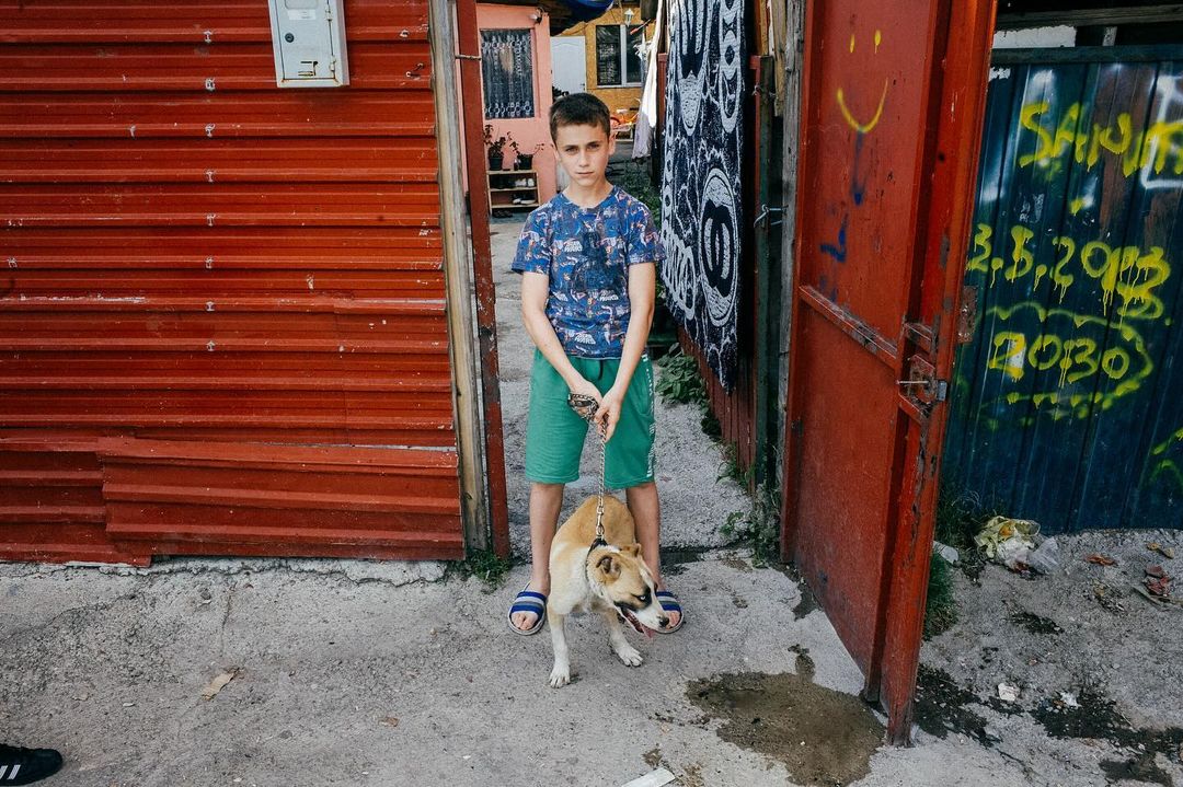 A young boy stands in a doorway with his dog on a leash in a somewhat impoverished urban area, with textured red and graffiti-covered walls.