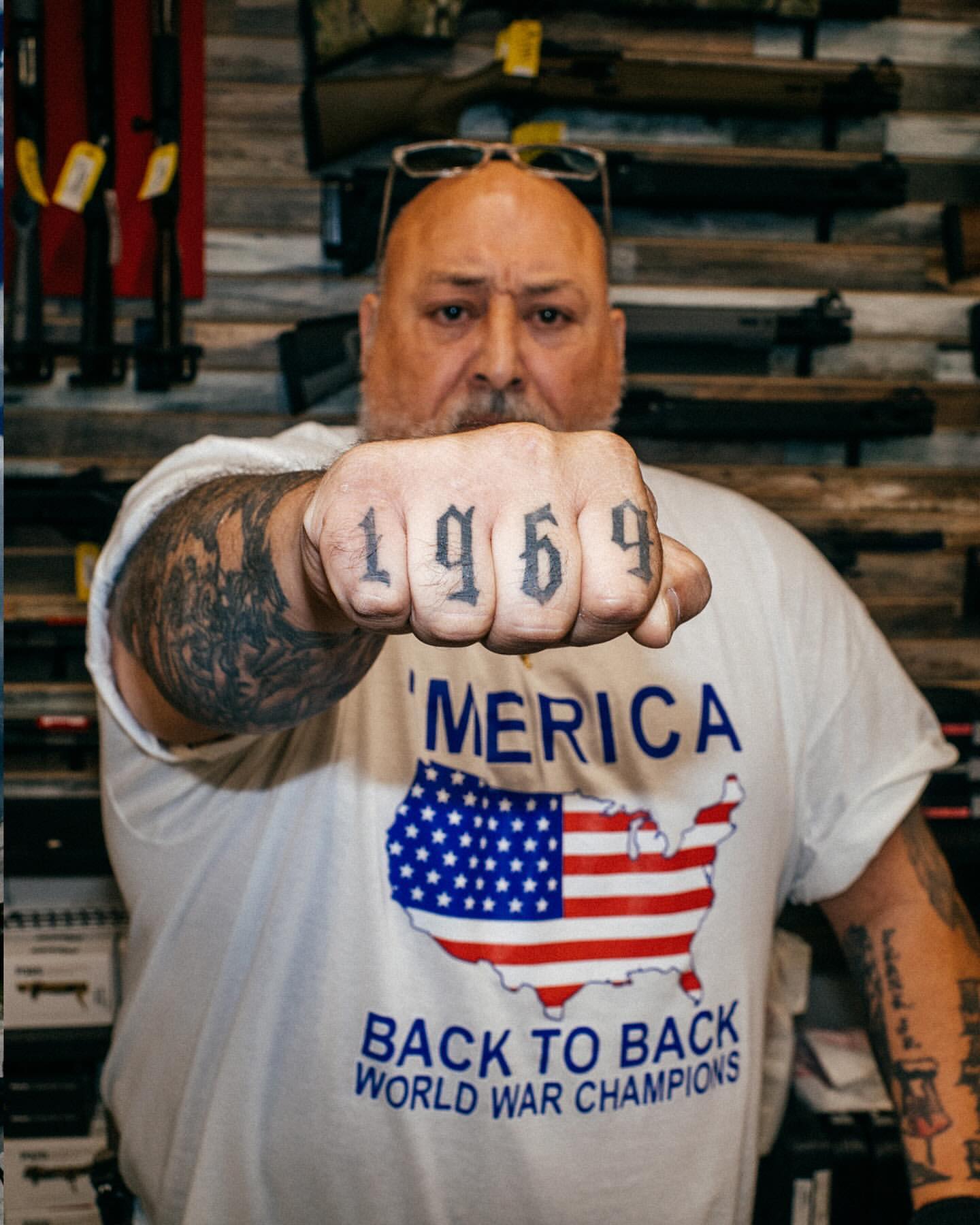 Man displays '1969' tattoo on knuckles, wearing a 'Back to Back World War Champions' shirt, standing in an indoor shop environment.