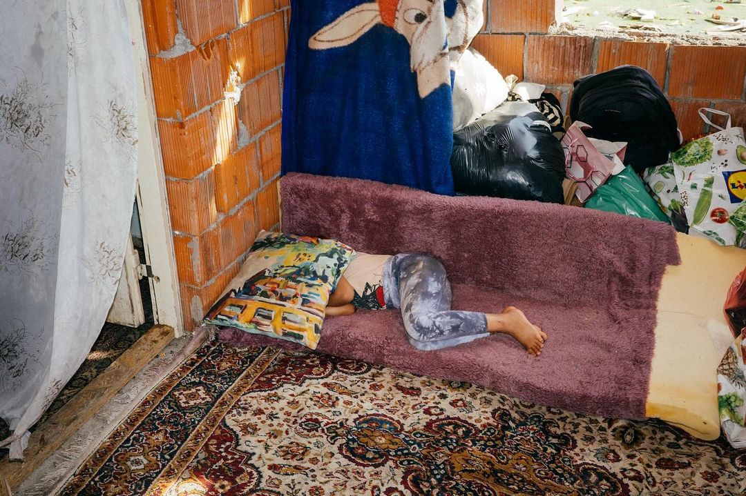 Person resting on purple sofa with patterned pillow in cluttered room, featuring patterned rug, exposed brick, and draped fabrics.