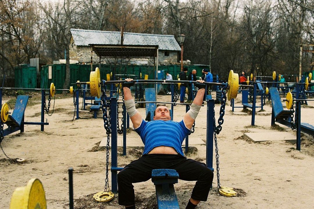 Adult male performs bench press with barbell in outdoor gym; weightlifting in a public workout park with exercise equipment.