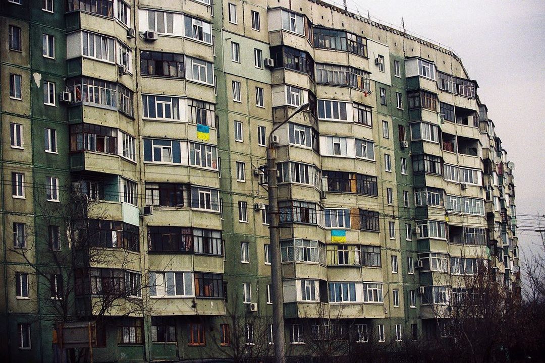Tall apartment building facade with balconies and windows, and Ukrainian flags hanging from some of the apartments.