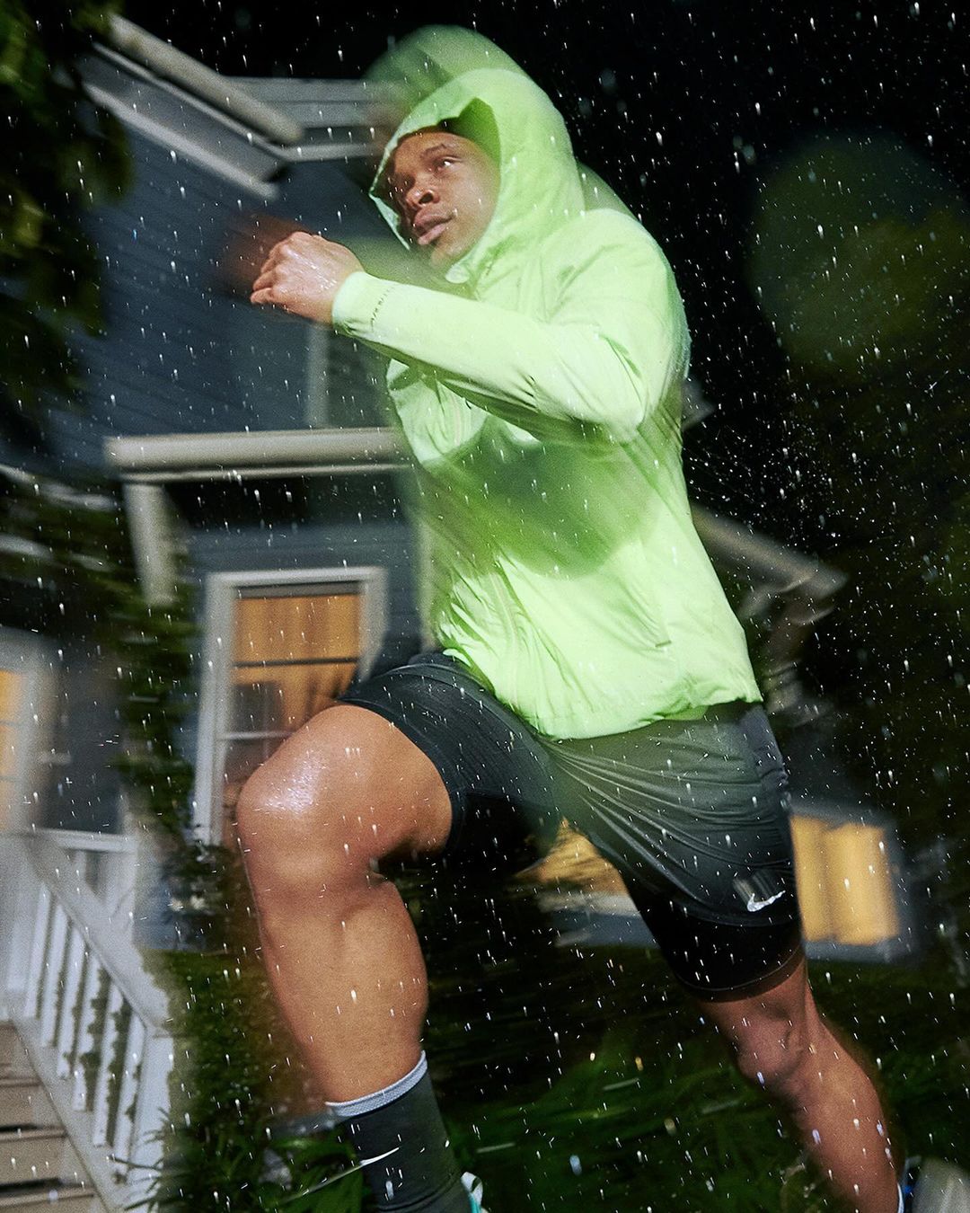 Athletic man in a hooded neon green jacket runs on the sidewalk in the rain at night, showcasing fitness and determination.