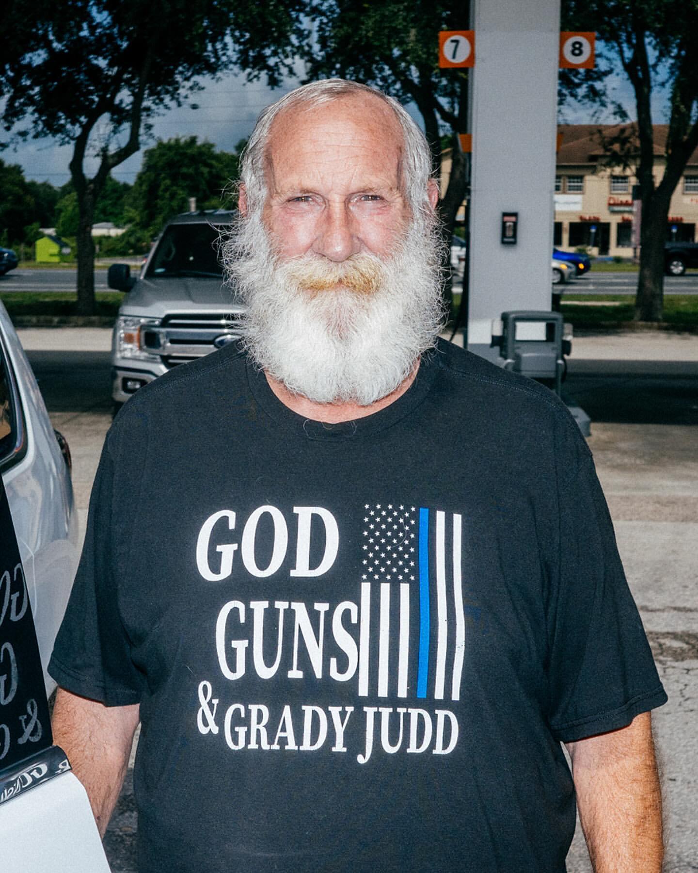 Man with long white beard stands in front of gas station wearing a black t-shirt with text and flag graphic. Outdoors.