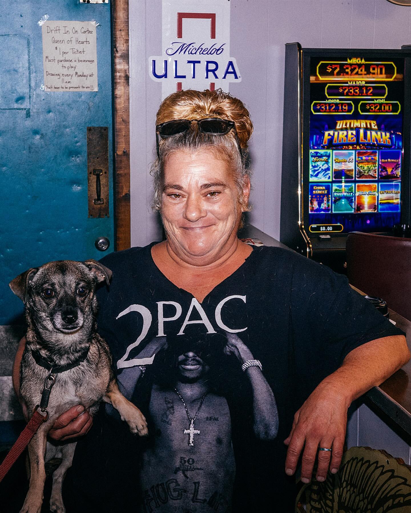 Adult woman holds a small dog in an indoor bar setting with a Michelob Ultra sign and slot machine visible in the background.