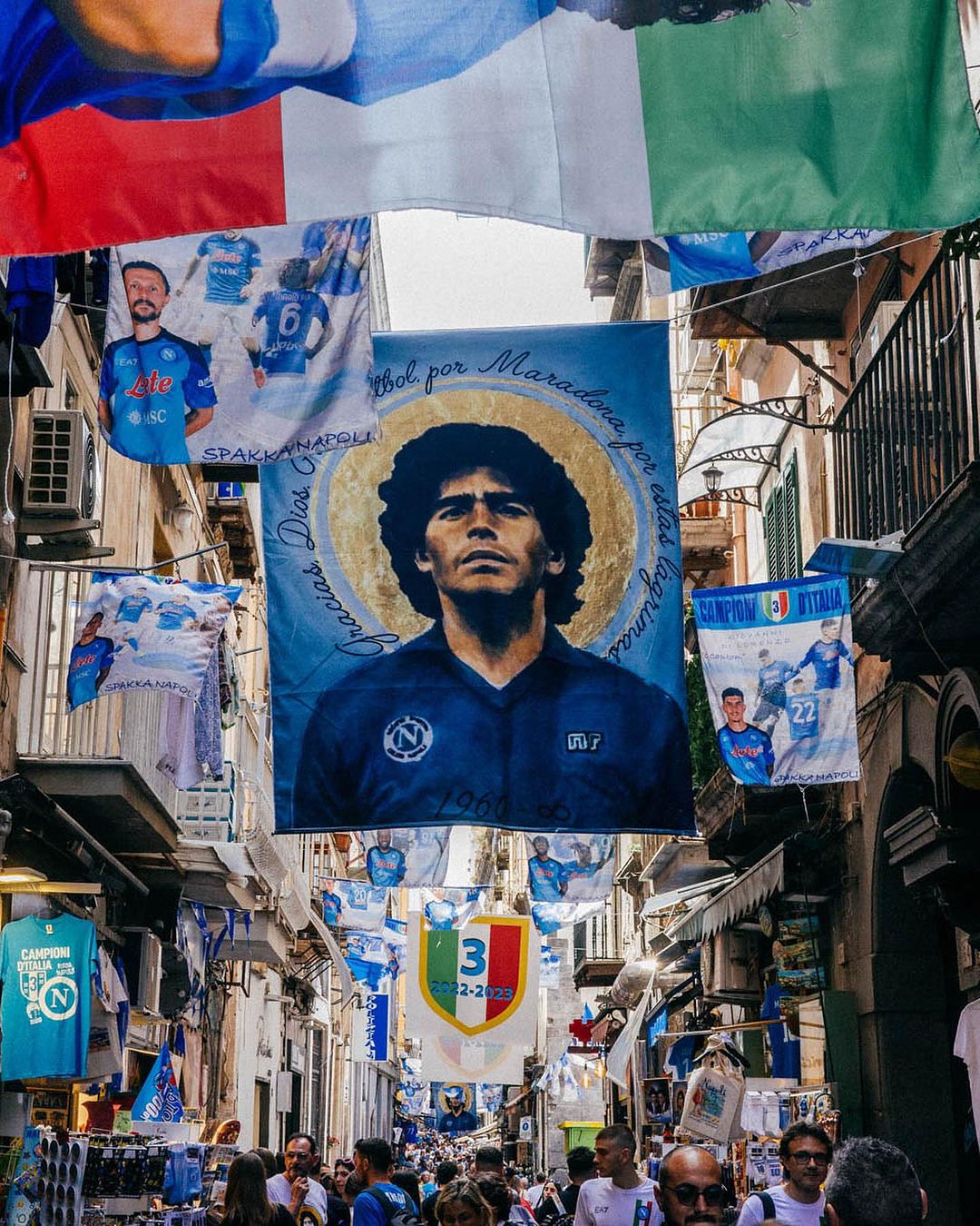Street in Naples, Italy decorated with flags and banners, including a large banner with an image of soccer player Diego Maradona.
