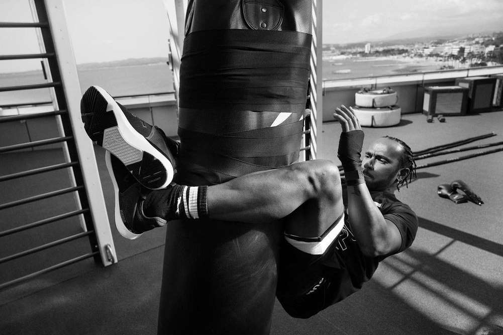 Black and white shot of an adult male doing sit-ups on a punching bag outdoors on a rooftop with a city and ocean in the distance.