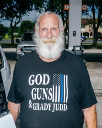 Elderly man with a long white beard wearing a black graphic t-shirt standing outdoors near a service station