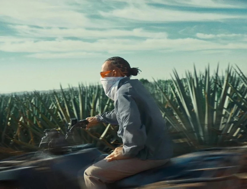 Person riding an ATV on a dirt path, wearing a light grey jacket and face covering, with an agave field and cloudy sky in the background.